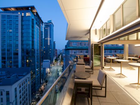 A modern cityscape at dusk features a rooftop terrace with seating, overlooking illuminated buildings and a street below.
