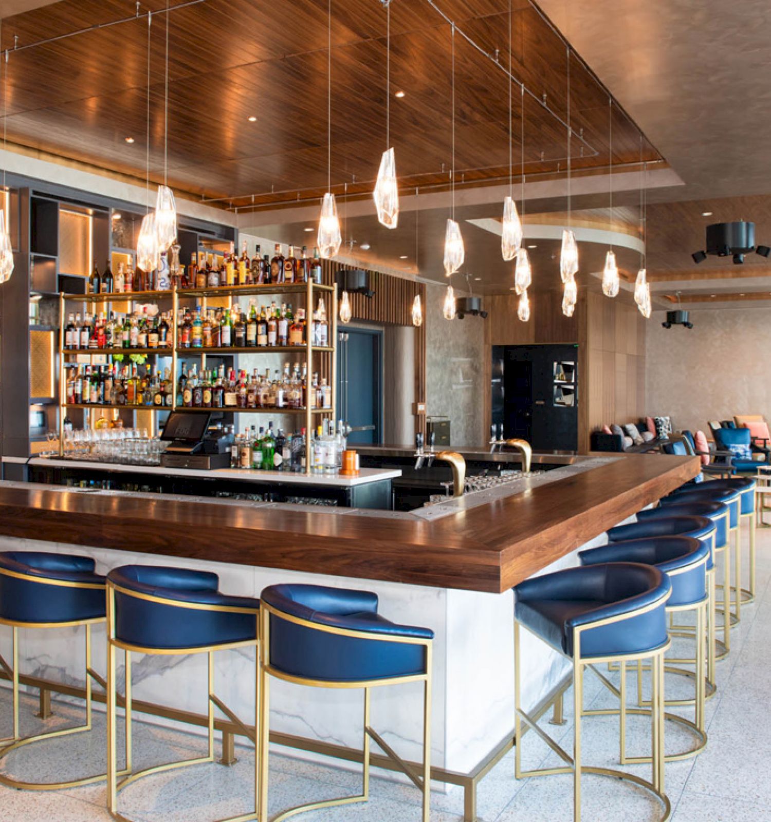 A modern bar with blue chairs, hanging lights, and a fully stocked shelf. The space is stylish, with a cozy seating area to the right.