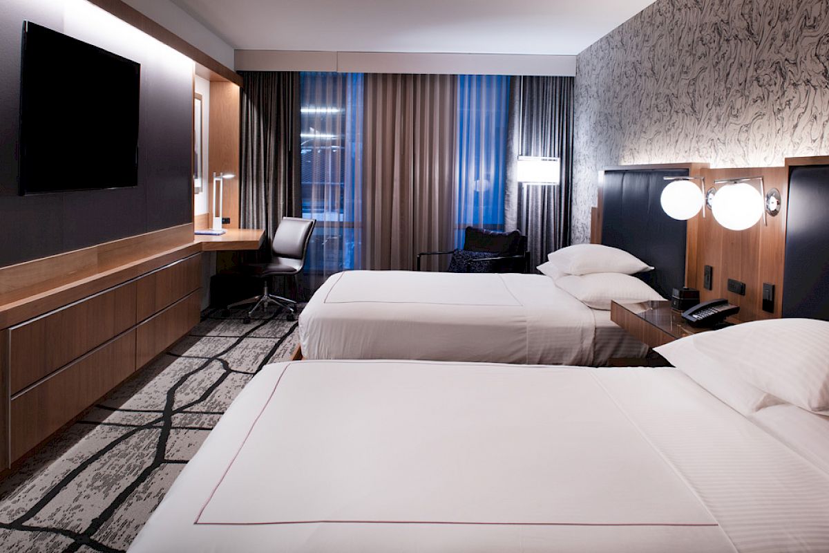 A modern hotel room with two beds, a TV, desk, and lamps. Décor features wood and soft lighting, with abstract patterns on the carpet and wall.