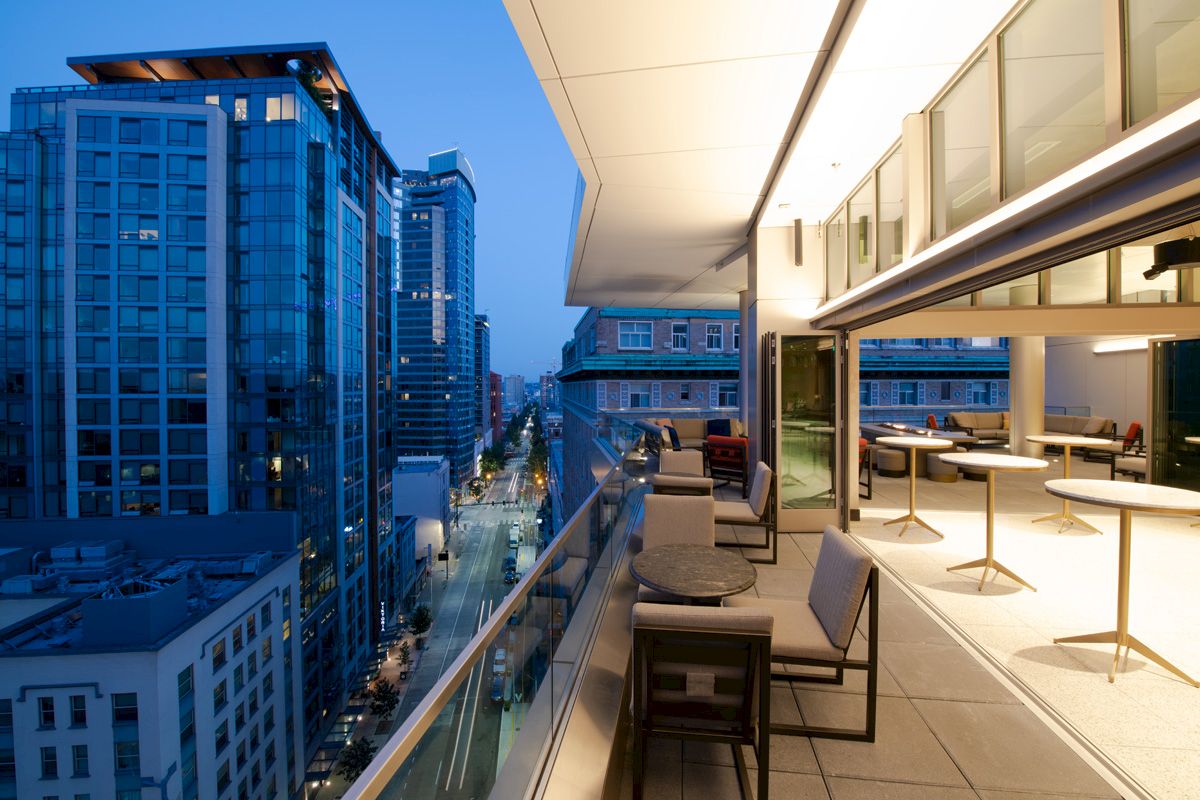 A modern city view from a balcony at dusk, with buildings, a street below, and outdoor seating on the terrace.