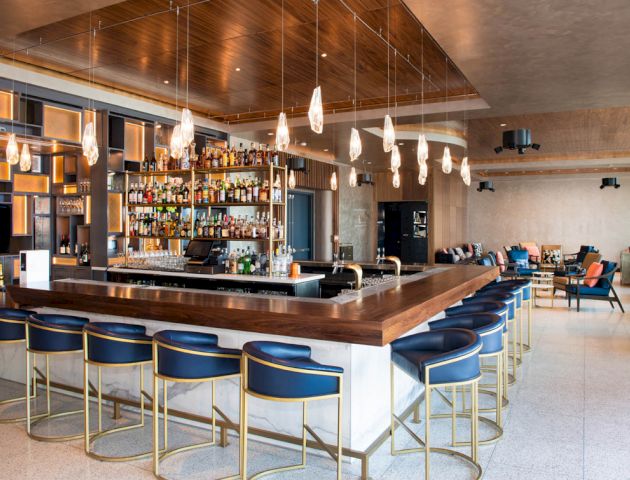 The image shows a modern bar with a wooden countertop, stylish seating, hanging lights, and an assortment of bottles displayed on shelves.
