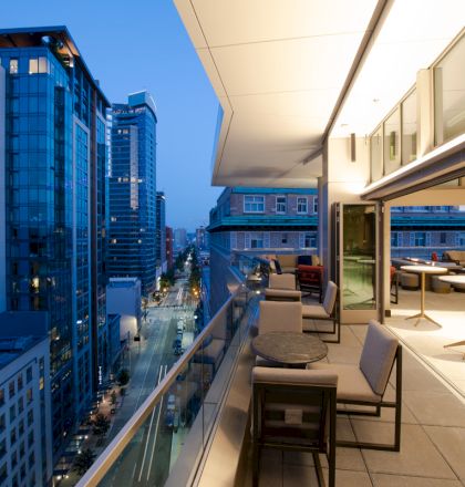 A cityscape at dusk with high-rise buildings and a modern, open-air balcony area with tables and seating, overlooking the street below.