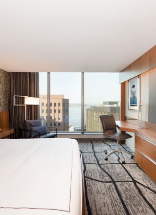 A modern hotel room with a bed, desk, chair, large TV, and window view of city buildings and water.