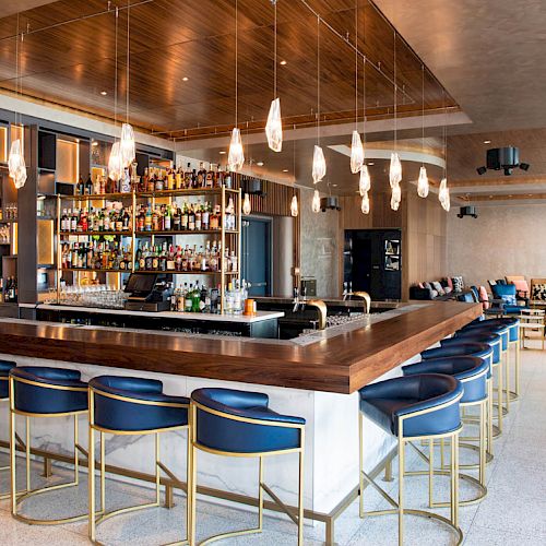 A modern bar with blue stools, wooden accents, and hanging lights. Bottles line the shelves, and there's a cozy seating area in the back.