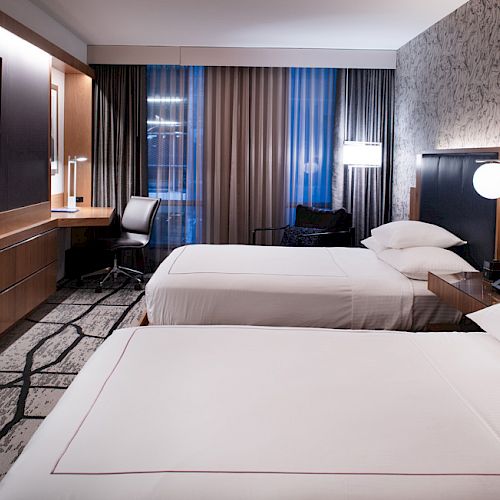 The image shows a modern hotel room with two beds, a TV, desk, chair, and stylish decor, featuring a patterned wallpaper.