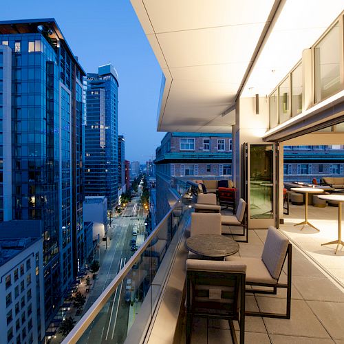 A modern rooftop terrace with tables and chairs overlooks a cityscape featuring tall buildings and a street at dusk.