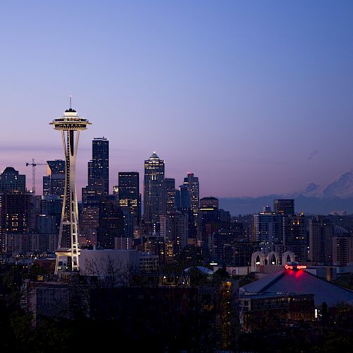 Seattle skyline at dusk with the Space Needle and Mount Rainier in the background, under a clear blue-purple sky.