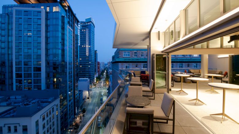 A cityscape view from a modern balcony at dusk, featuring buildings, a street, and outdoor seating with tables and chairs.