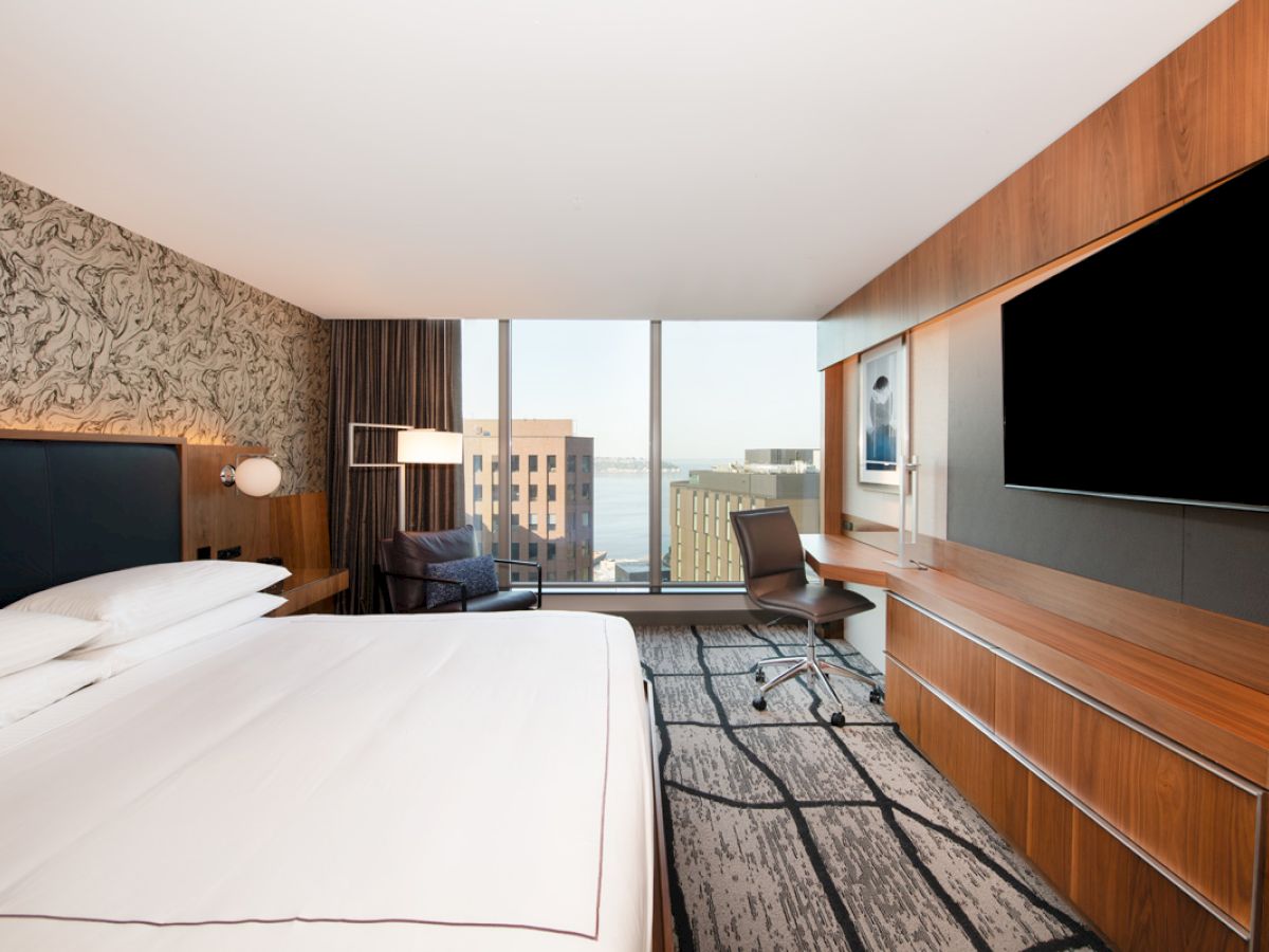 A modern hotel room with a large bed, wall-mounted TV, desk, chair, and a view of buildings through a window.