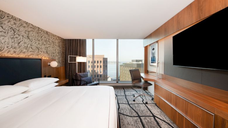 A modern hotel room with a large bed, wall-mounted TV, desk, chair, and a view of buildings through a window.