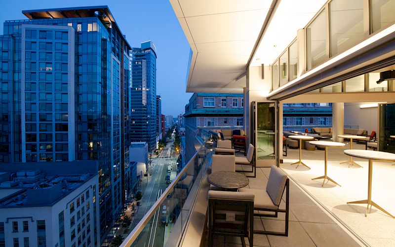 A modern balcony view featuring comfortable seating, tables, and cityscape with buildings at twilight.