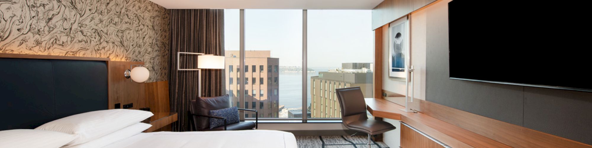 A modern hotel room with a large bed, desk, chair, and a flat-screen TV, overlooking city buildings through a large window.